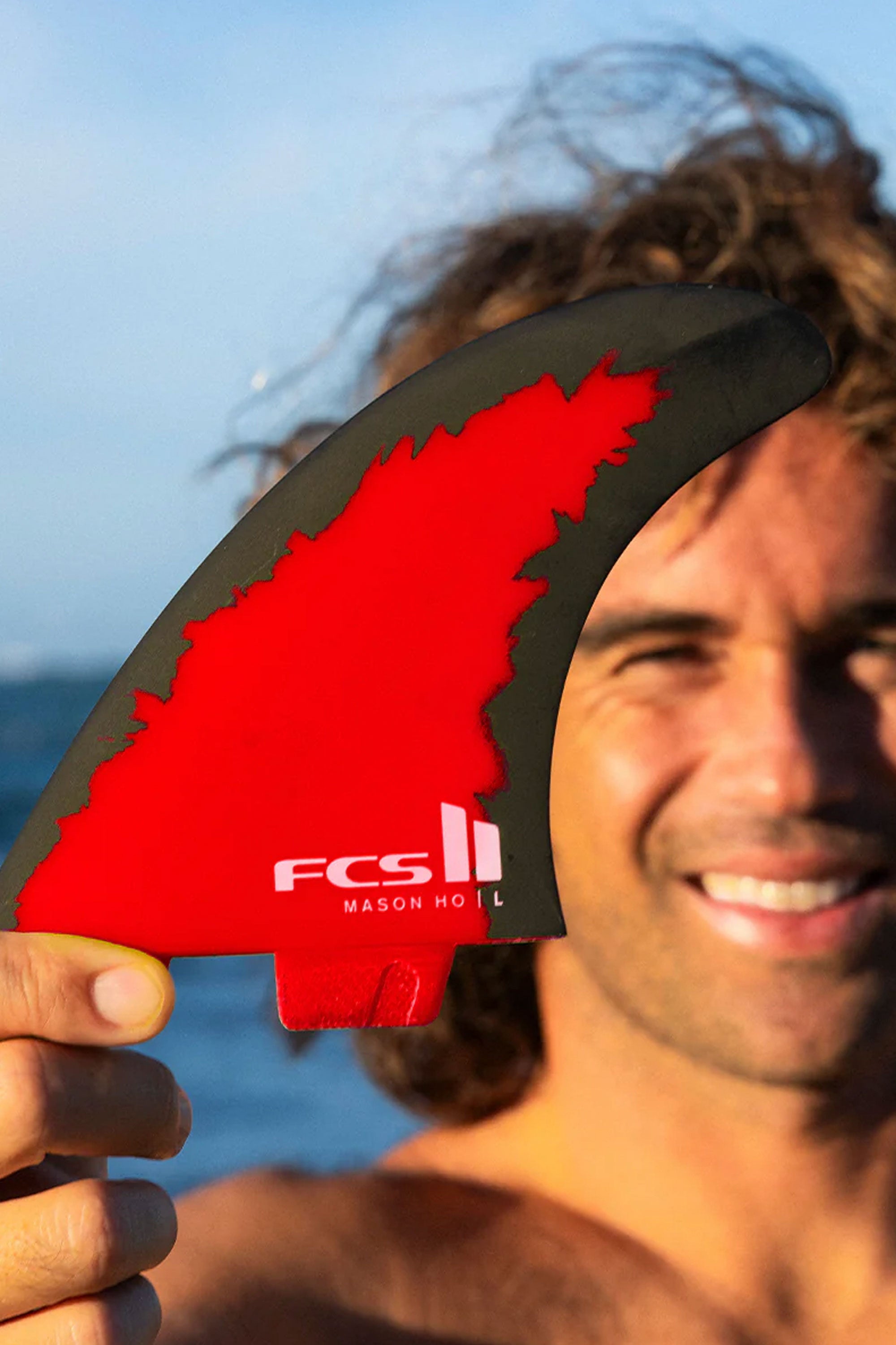 Man holding a red FCS II surfboard fin against a blue sky and ocean background