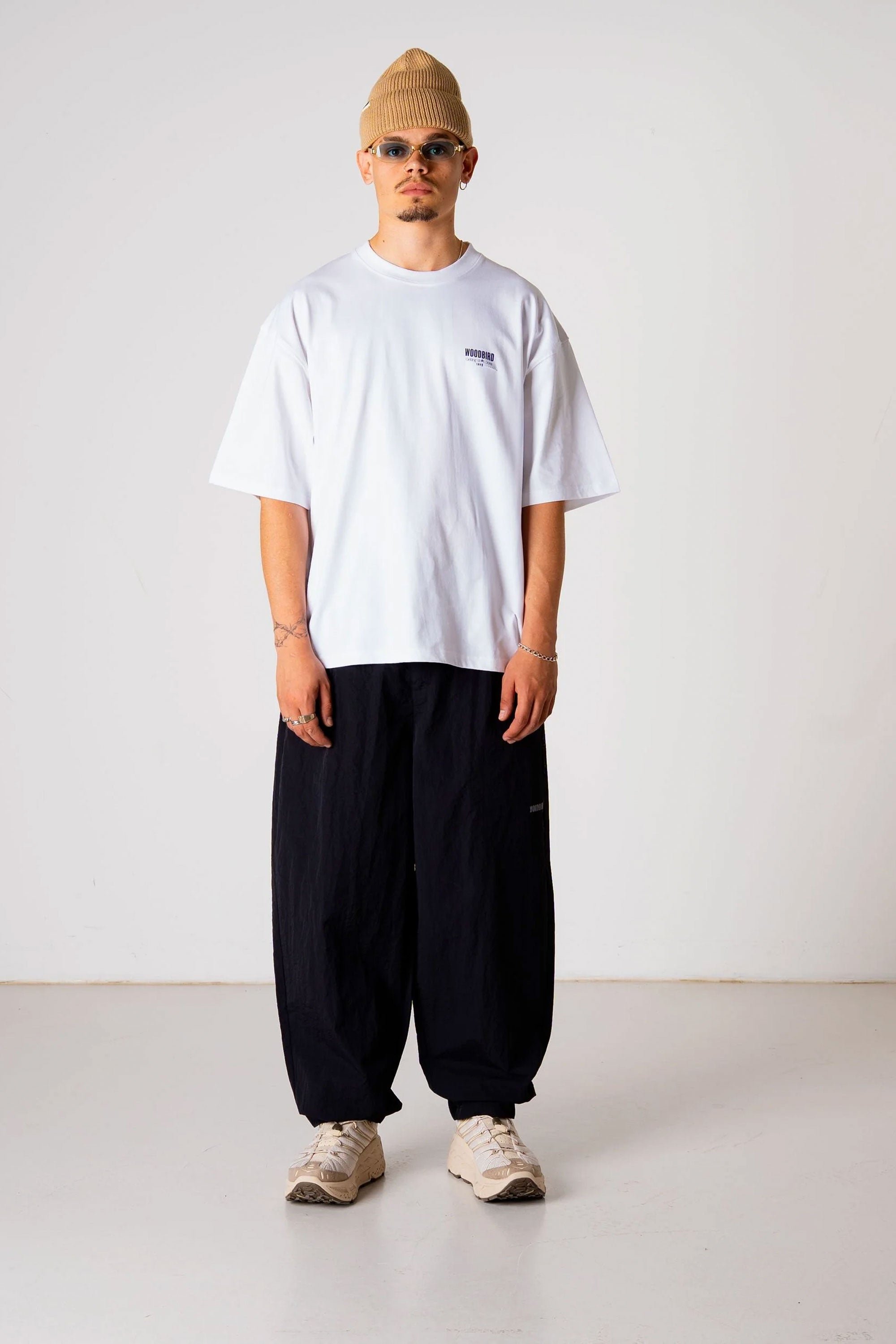 Person wearing a white t-shirt, black pants, and beige beanie standing against a plain background